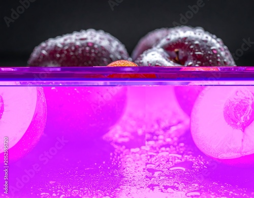 A halved plum reveals its vibrant orange interior with a dark center, surrounded by two whole plums, all adorned with water droplets on a dark surface