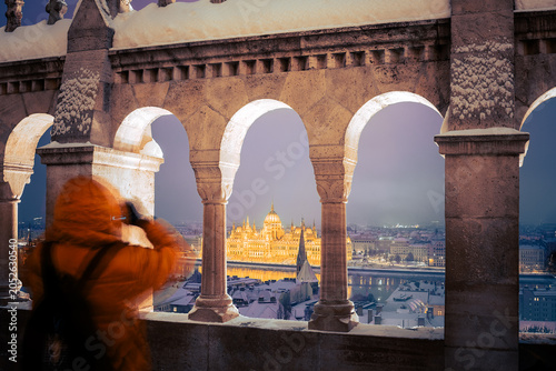 Photographer in winter jacket taking photos of the illuminated Hungarian Parliament Building through the arches of Fisherman's Bastion at dusk, Budapest, Hungary