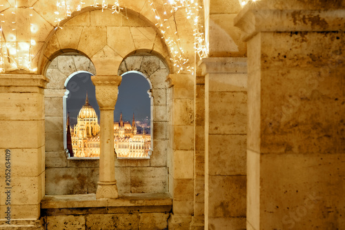 Illuminated Hungarian Parliament dome framed by stone window of Fisherman's Bastion decorated with Christmas lights at night, Budapest, Hungary