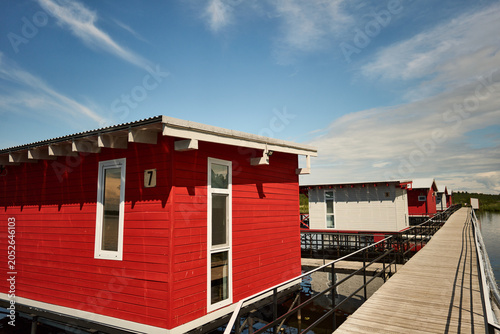 Photos Red-painted wooden boathouses and a white boathouse built on stilts over a calm lake, connected by a narrow boardwalk and metal railings