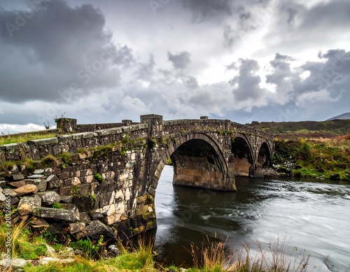A historic stone bridge spans a serene river under a stormy sky