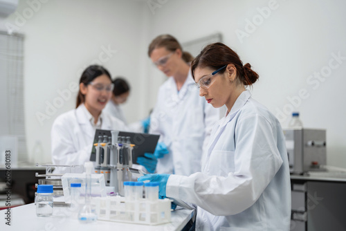 Tableau sur toile Professional female medical scientist reviewing clinical trial data on a digital tablet with diverse teamwork colleagues working in the background of a modern commercial pharmaceutical lab