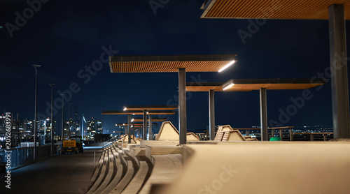 Photos Modern waterfront promenade at night with illuminated wooden canopy structures and concrete benches