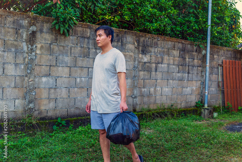 Картината върху платно Middle-Aged Asian Man Carrying Full Black Trash Bag While Walking by Concrete Bl