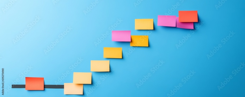 Fototapeta premium Multicolored sticky notes arranged diagonally upward from a dark baseline on a bright blue background, illustrating progress, growth and achieving milestones. Planning, Strategy
