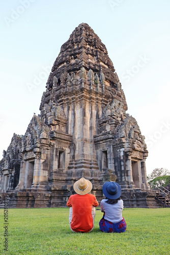 Tourist couple sitting on the grass, admiring the majestic Phimai ancient stone castle, Phimai historical park, Nakhon Ratchasima, Thailand