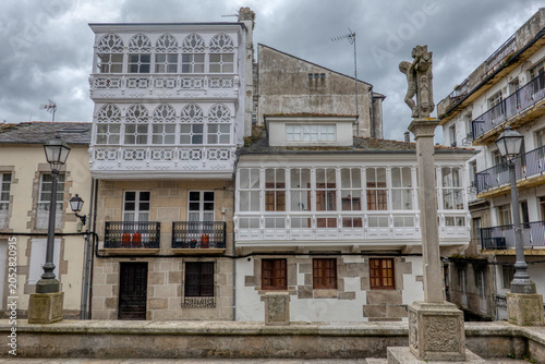 Traditional white glass balconies and an ancient stone cross in the historic center of Viveiro, Galicia, Spain under a cloudy sky.
