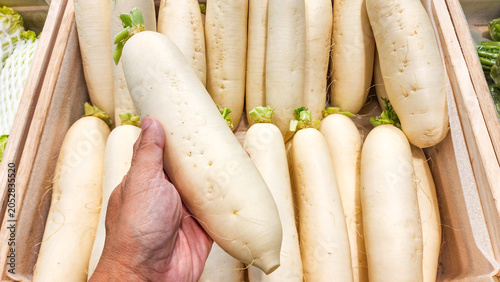A hand holding a large, fresh white daikon radish over a wooden crate filled with radishes at a supermarket grocery display counter.
