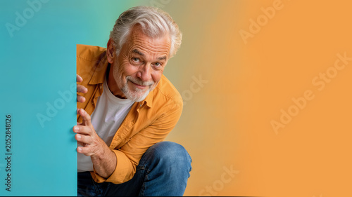 Smiling senior man appears from behind a colored panel with a friendly gesture, ideal for presentation, surprise, or advertising concepts. Senior citizen peeking out from behind a panel with a friend