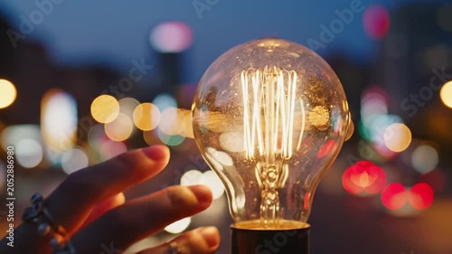 Hand with rings holding glowing vintage light bulb against city bokeh lights at night