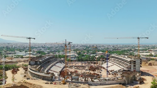 High aerial view of a massive stadium construction site in an urban area with several tall tower cranes working on the concrete structure on a sunny day with blue sky in the background