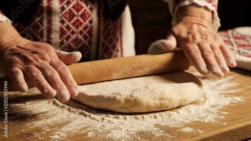 Hands roll dough on a floured wooden table in warm kitchen light, traditional embroidered clothing suggests heritage cooking, calm homely mood for baking