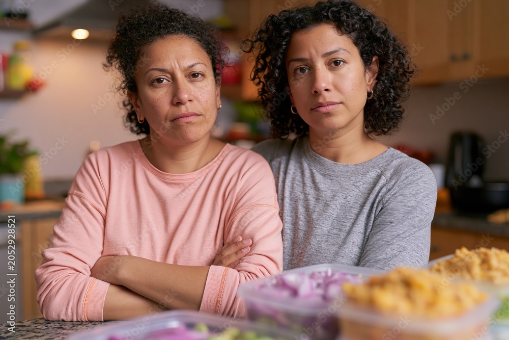 Fototapeta premium Two women with curly hair standing close together in a kitchen, arms crossed, with food containers on the counter and a modern kitchen setting in the background