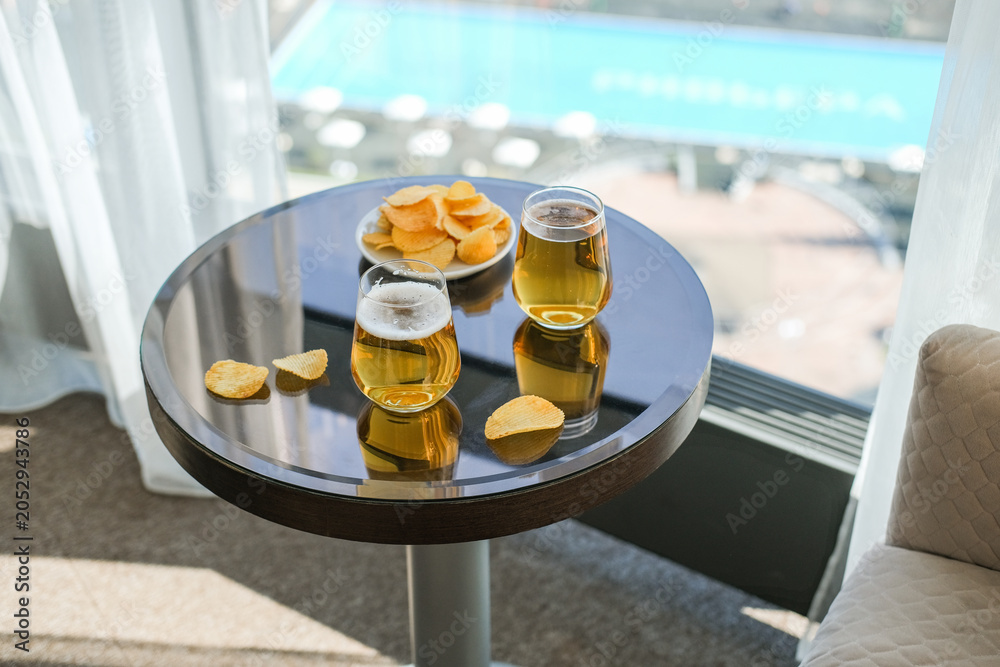 Fototapeta premium Two glasses of cold beer and potato chips on reflective table overlooking a resort pool. Bright sunny concept of travel, leisure and summer vacation relaxation in hotel room