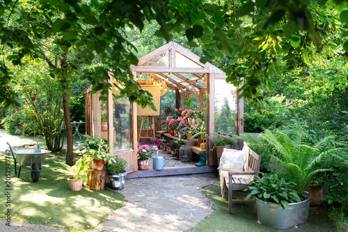 Cozy exterior wooden glass greenhouse in a backyard garden filled with potted plants and flowers. Beautiful outdoor relaxation area with a wooden bench, pillows, stone path, and lush green trees.