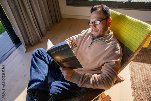 Asian man reclining in chair at home near glass door, wearing glasses and reading ENDLESS SUNSETS