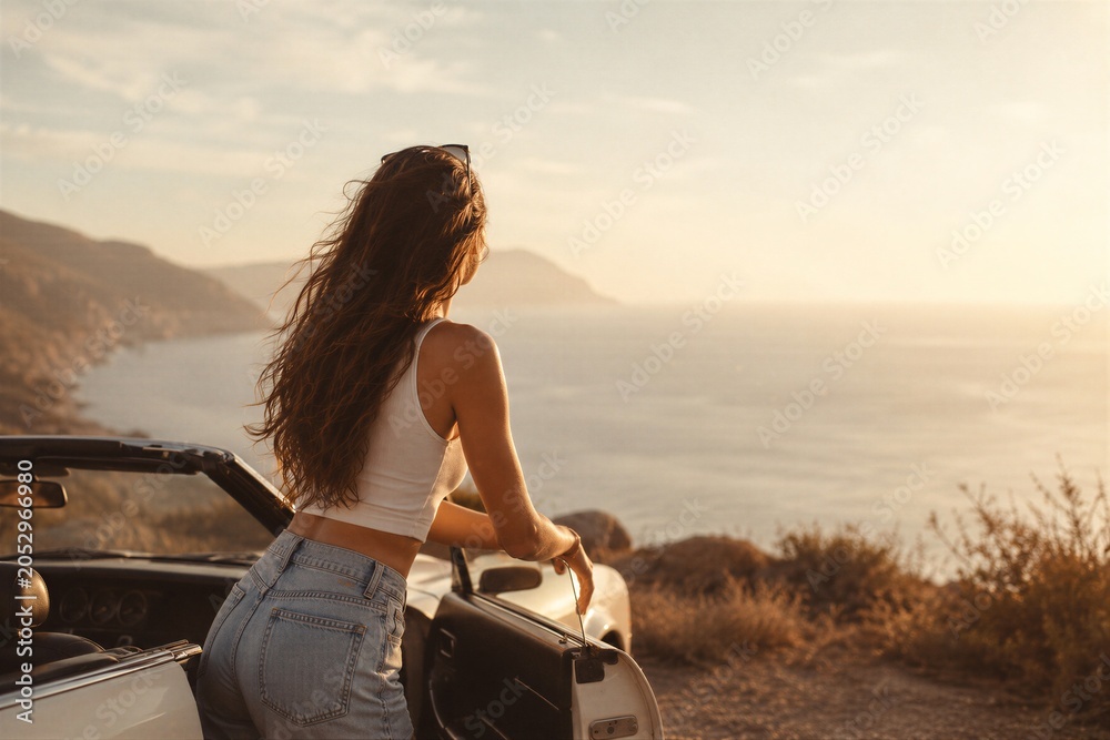 Fototapeta premium Woman standing by convertible car looking at seaside sunset. Female traveler enjoying view of ocean during road trip. Summer vacation, freedom, travel concept and coastal adventure