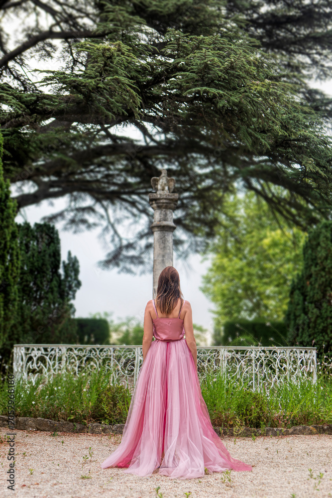 Fototapeta premium Woman in elegant pink gown standing in a historical garden from behind.