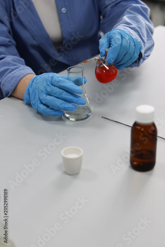 Scientist wearing protective gloves pours chemical solution into laboratory flask during scientific experiment. Biotechnology research, chemical analysis and laboratory testing concept.