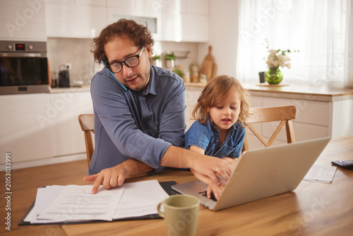 Fotografie A father helps his child with a laptop while surrounded by documents on a table in their cozy kitchen