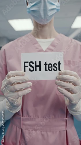 Hands in a medical gown and gloves hold a sign with the text FSH test in a laboratory