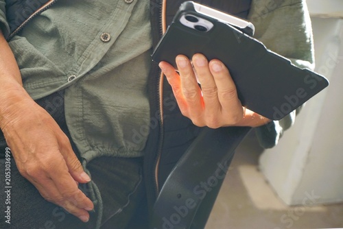 Sitzende schlanke ältere Frau auf Bürostuhl mit grauer Jeanshose und grüner Bluse hält ihr Handy in der Hand und liest in Raum bei Sonne am Abend im Frühling