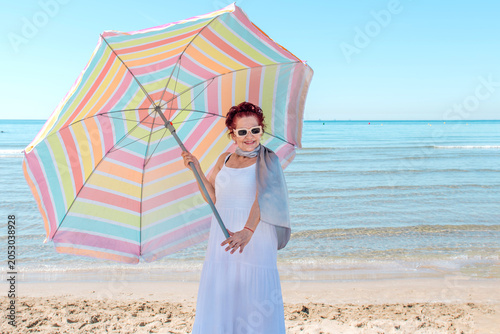 A senior woman in fashionable sunglasses and summer senior wear with a colorful umbrella enjoys relaxing on the beach by the sea on a sunny day. tourism, summer accessories. Senior wear in the heat