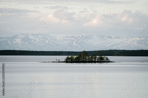 Remote tree-covered island on calm lake with snow-capped mountains