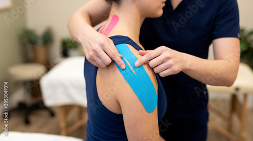 Physiotherapist Applying Blue Kinesio Tape on Woman Shoulder. Kinesiology Taping Treatment for Muscle Pain Relief, Sports Injury Rehabilitation and Physical Therapy in Clinic.