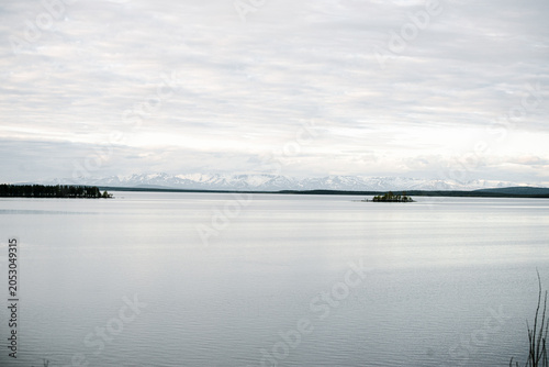Calm lake with distant tree line and snowy mountains under overcast sky