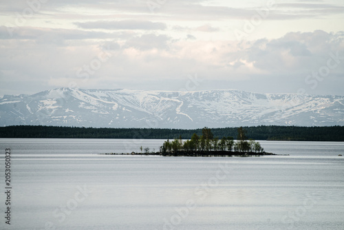 Small tree-covered island in a calm northern lake with distant snow-capped mountains