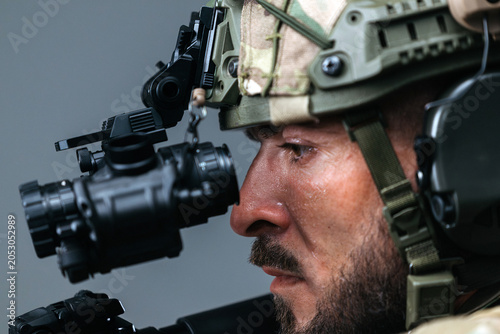 Armed special forces operator in tactical gear with weapon raised, equipped with night vision device, focused expression, and camouflage helmet against a neutral background