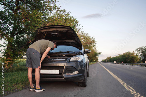 Man examining engine under open hood of broken car parked on highway shoulder, roadside vehicle failure during journey, transportation problem concept, automotive diagnostics, emergency 