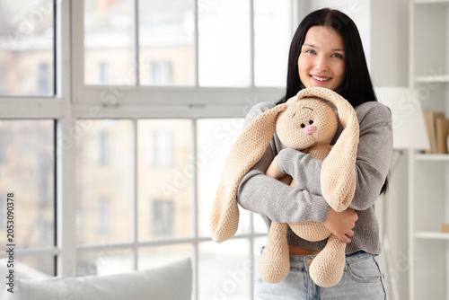 Beautiful young woman hugging toy bunny near window at home