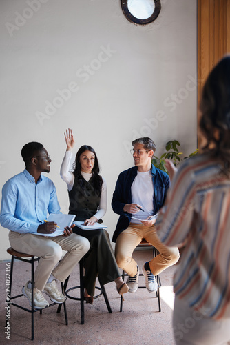 Diverse business professionals on stools actively participating in a workshop—listening to a speaker, taking notes, raising hands, and engaging in discussion and learning