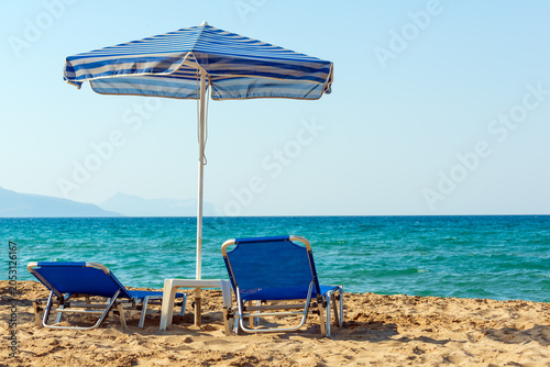 Two blue sun beds under blue stripped umbrella on sandy Petres beach near Rethymno in Crete, Greece