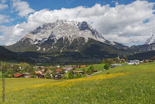 Blick über blühende Wiesen, das Dorf Lermoos und auf das Zugspitzmassiv mit Wolken, dazwischen im Schatten Ehrwald. Tiroler Zugspitz Arena, Tirol, Österreich.