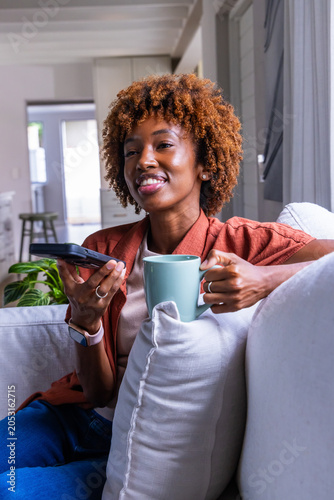 African American adult woman sitting at home on sofa holding teal mug, black remote, rust shirt
