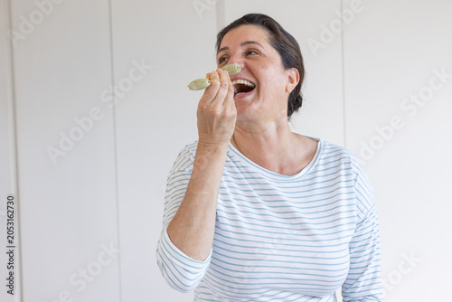 Принти на полотні Mature woman laughing, lifting small spoon toward mouth at home wearing white st