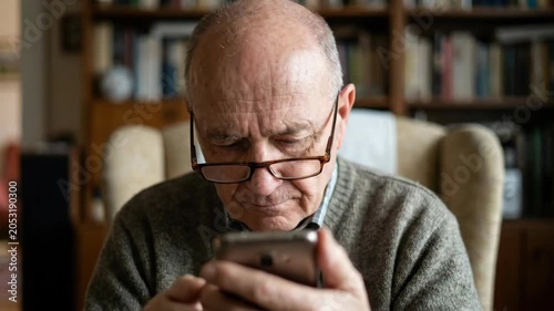 Elderly man using smartphone at home with glasses, focused expression in living room, senior home reading scene