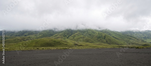 A serene, wide-angle landscape photograph capturing the dramatic, deep valleys and jagged mountain slopes of the Bromo-Tengger-Semeru region under thick, layered fog. The soft, diffuse light filters t
