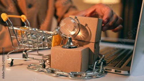 Foto A businessperson arranges boxes beside a miniature globe and a shopping cart wrapped in chains illustrating small entrepreneurs’ ongoing struggle against monopolistic powers in the global market