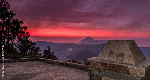 A breathtaking high-altitude panoramic view at dawn or dusk, looking over a vast, layered mountain range under a dramatically vibrant crimson and magenta sky with a bright orange light source at the h