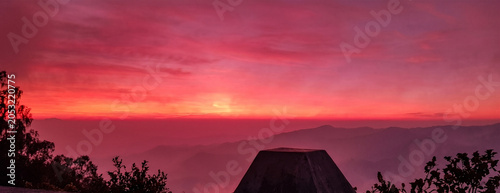 A breathtaking high-altitude panoramic view at dawn or dusk, looking over a vast, layered mountain range under a dramatically vibrant crimson and magenta sky with a bright orange light source at the h
