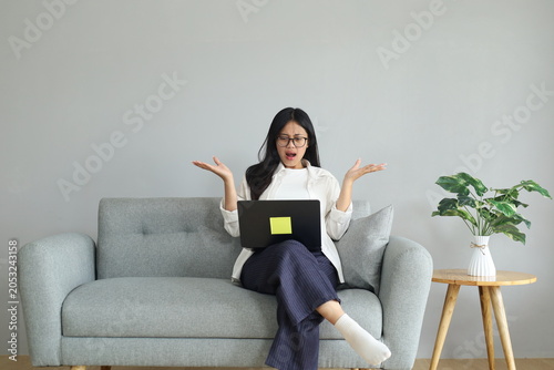 Картината върху платно A young working woman sits on a sofa in the living room, staring anxiously at a laptop screen, reacting to bad and unexpected news, frustrated and angry, shrugging her shoulders and hands