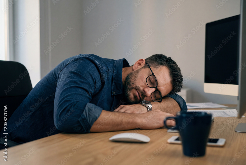 Fototapeta premium Man with a beard and glasses sleeping on his arms at a wooden office desk