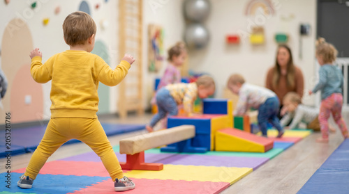 Toddler in yellow tracksuit, standing on colorful foam mats, watching children play, indoor gym daycare center, lifestyle