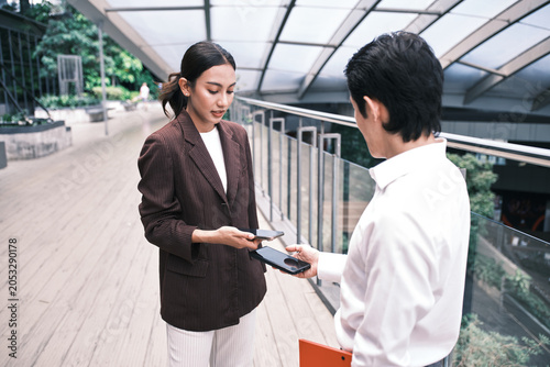 Young Asian smart businesspeople exchange digital contact information with NFC QR code, wireless mobile phone app technology, contactless startup collaboration at outdoor workplace, modern lifestyle.