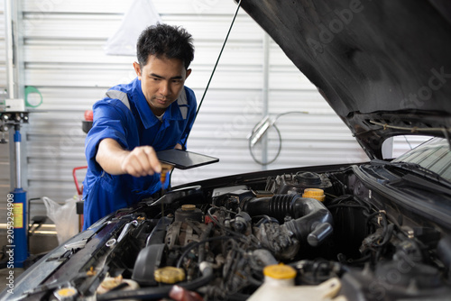 Two mechanics examine a car engine together. One holds a clipboard, the other a laptop. They focus on the engine's components closely. Their expressions show concentration and teamwork
