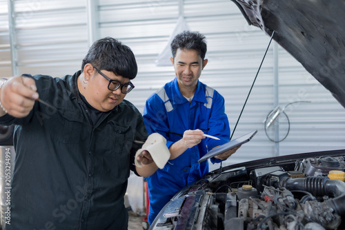 Two mechanics examine a car engine together. One holds a clipboard, the other a laptop. They focus on the engine's components closely. Their expressions show concentration and teamwork